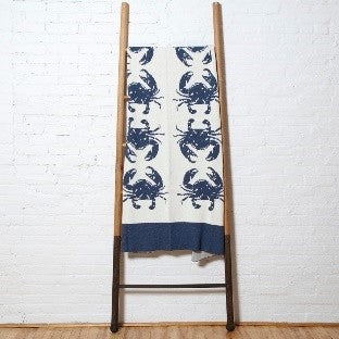 A blue and white cotton throw blanket by In2Green with blue crab patterns displayed on a wooden ladder.