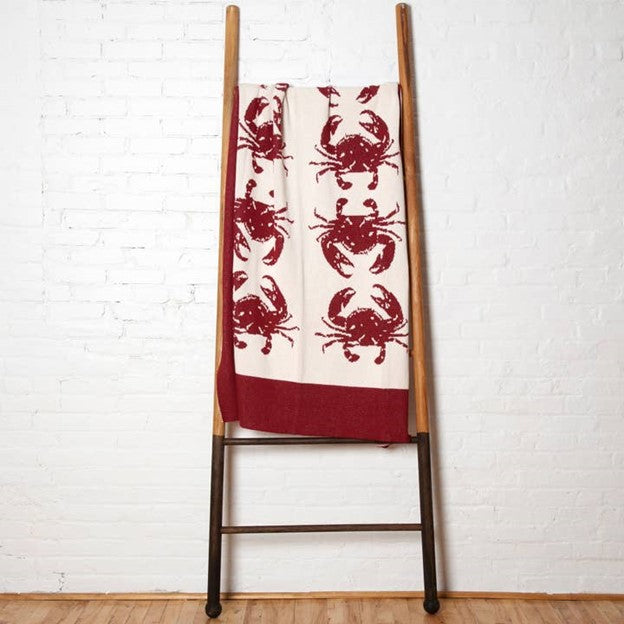A red and white cotton throw blanket by In2Green with crab patterns displayed on a wooden ladder.