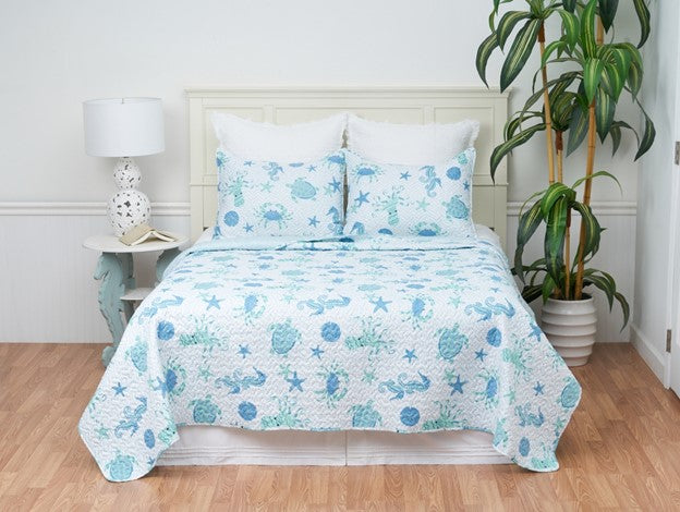 A bed with a blue quilt set featuring starfish and coral patterns, with two shams, placed in a room with a nightstand and a plant.