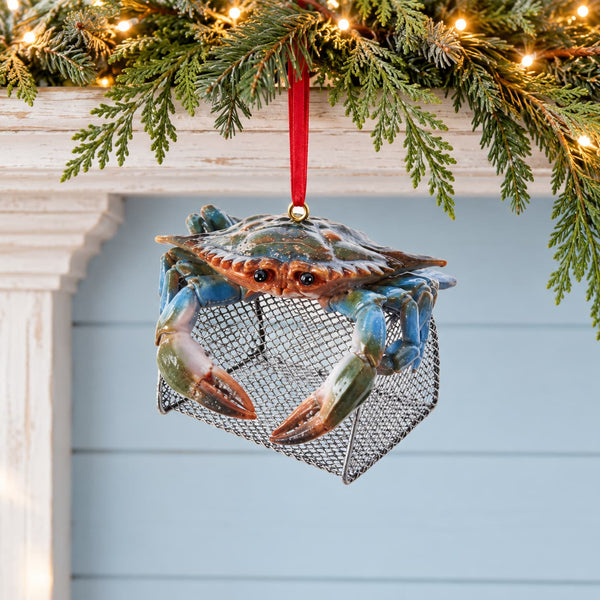 A decorative hanging ornament shaped like a glass blue crab sitting inside a metal lobster trap, suspended by a red ribbon. The ornament is shown hanging from evergreen branches with warm string lights against a light blue paneled wall.