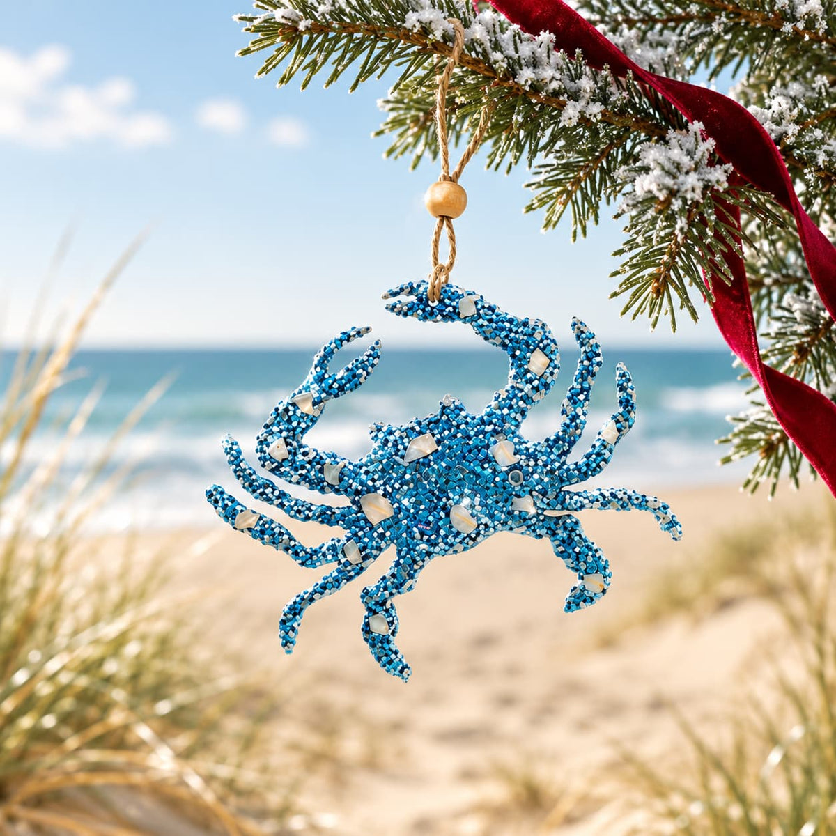 A glittery blue crab-shaped Christmas ornament hanging from a pine tree branch with a red ribbon, set against a blurred beach and ocean background.