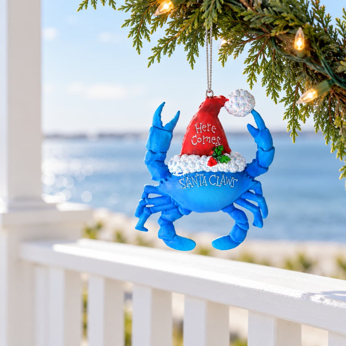 Blue Santa Claws crab Christmas ornament wearing a Santa hat with 'Here Comes Santa Claws' text hanging from a string on a branch overlooking the water