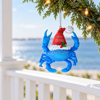 Blue Santa Claws crab Christmas ornament wearing a Santa hat with 'Here Comes Santa Claws' text hanging from a string on a branch overlooking the water