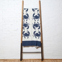 A blue and white cotton throw blanket by In2Green with blue crab patterns displayed on a wooden ladder.