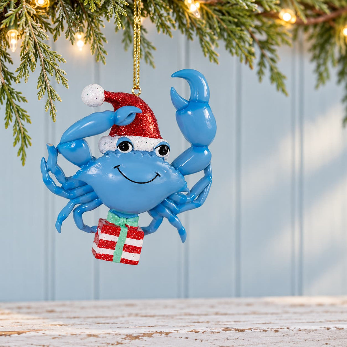 A whimsical blue crab Christmas ornament wearing a red sparkly Santa hat and holding a red and white gift box, suspended by a gold string
