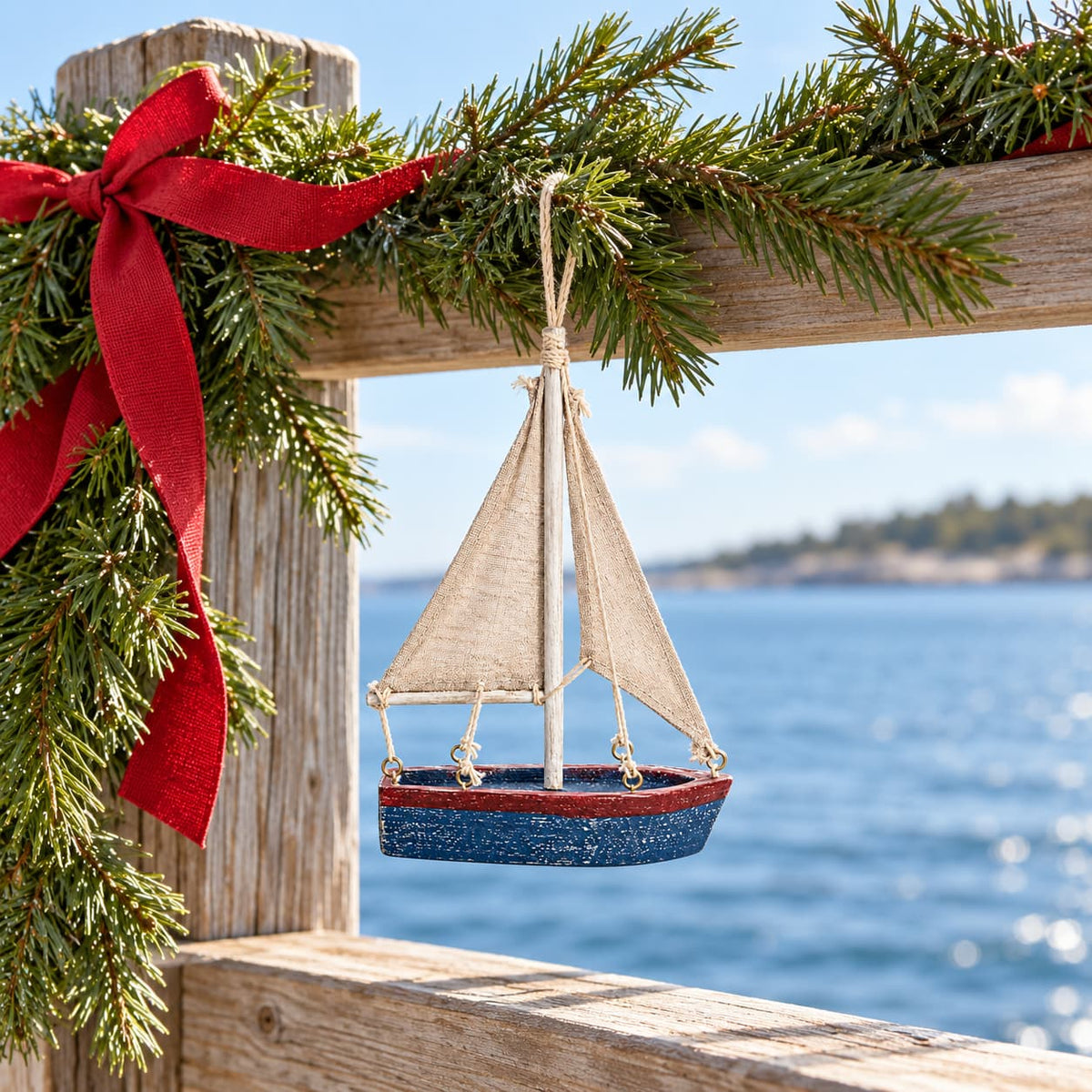 A sailboat ornament with a distressed blue hull, a red stripe, beige canvas sails, and a silver mast hanging from Garland over a bay.
