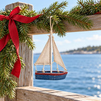 A sailboat ornament with a distressed blue hull, a red stripe, beige canvas sails, and a silver mast hanging from Garland over a bay.