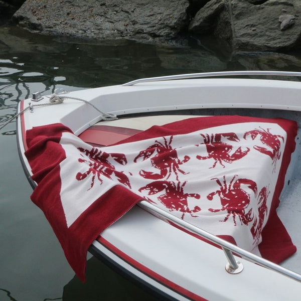A red and white cotton throw blanket by In2Green with crab patterns displayed on a wooden ladder.