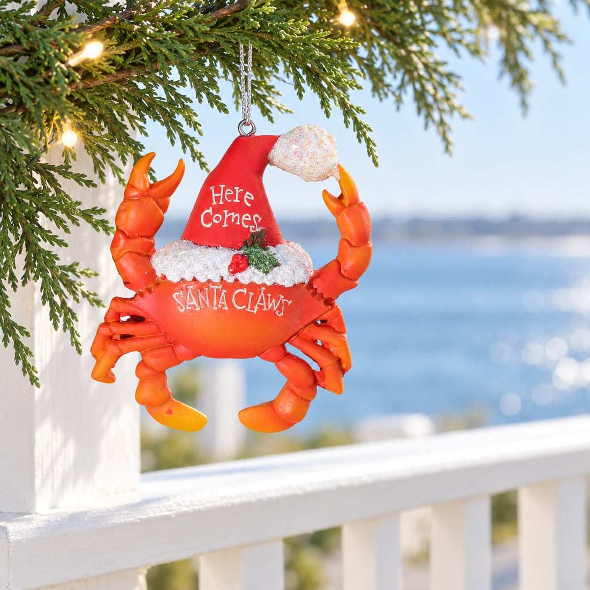 Santa Claws red ceramic Christmas Ornament with Santa hat hanging from Christmas branch overlooking water.