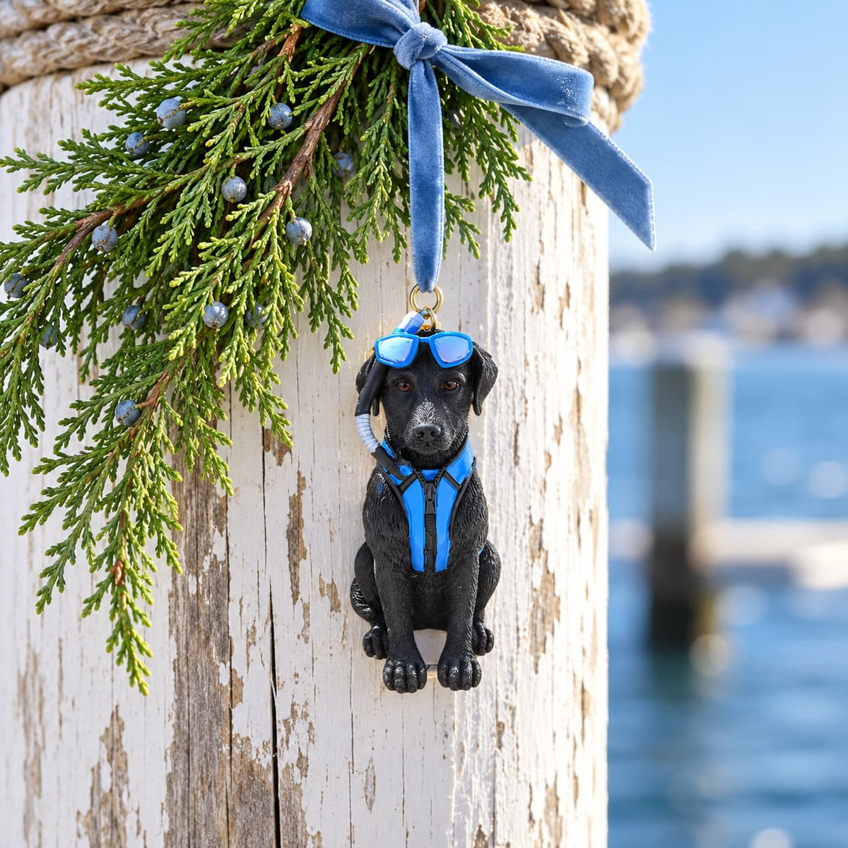 Black dog Christmas ornament with blue goggles and scuba gear hanging from pine branch on a dock