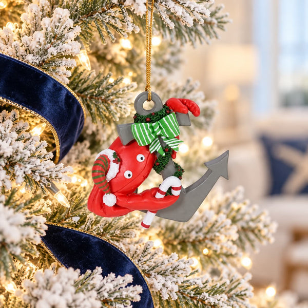 A Christmas ornament shaped like a red crab climbing an anchor, decorated with a green striped hat and red-and-white striped socks, hanging on a flocked Christmas tree with warm lights and a blue ribbon.
