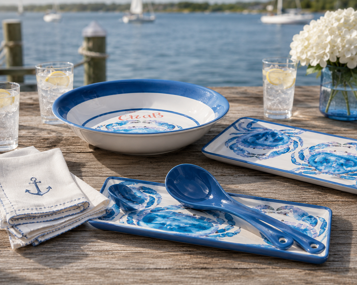Nautical-themed ceramic tableware set with blue and white crab illustrations, including a bowl, rectangular platters, and blue spoons, styled on a wooden table by the water with drinks and flowers.