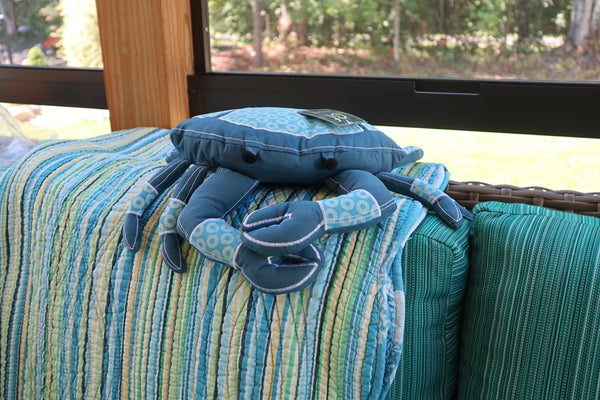 Small blue coastal crab shaped pillow with legs on a striped cushion with a window view in the background