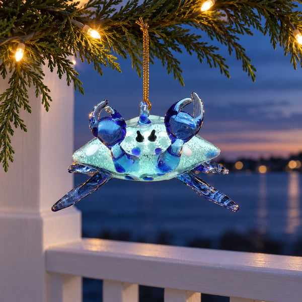 A blue and light blue glow in the dark glass crab ornament hanging from a gold string on a Christmas tree branch, with warm string lights and a waterfront sunset in the background.
