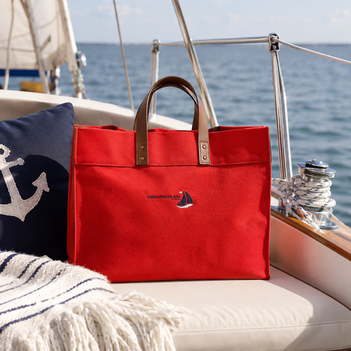 A red structured canvas tote bag with brown leather handles sits on a boat cushion, with the ocean and sailboat rigging in the background. 
