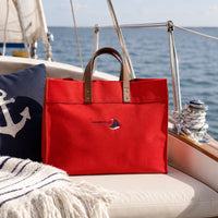 A red structured canvas tote bag with brown leather handles sits on a boat cushion, with the ocean and sailboat rigging in the background. 