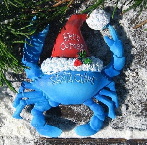 Blue Santa Claws crab Christmas ornament wearing a Santa hat with 'Here Comes Santa Claws' text on a sandy background.