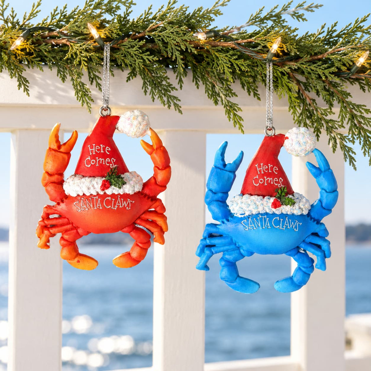 Blue and red Santa Claws crab Christmas ornament wearing a Santa hat with 'Here Comes Santa Claws' text hanging from a string on a branch overlooking the water