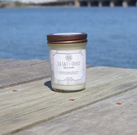 A jar candle labeled 'Sea Salt + Orchid Soy Candle' sits on a weathered wooden dock with a calm blue body of water in the background.