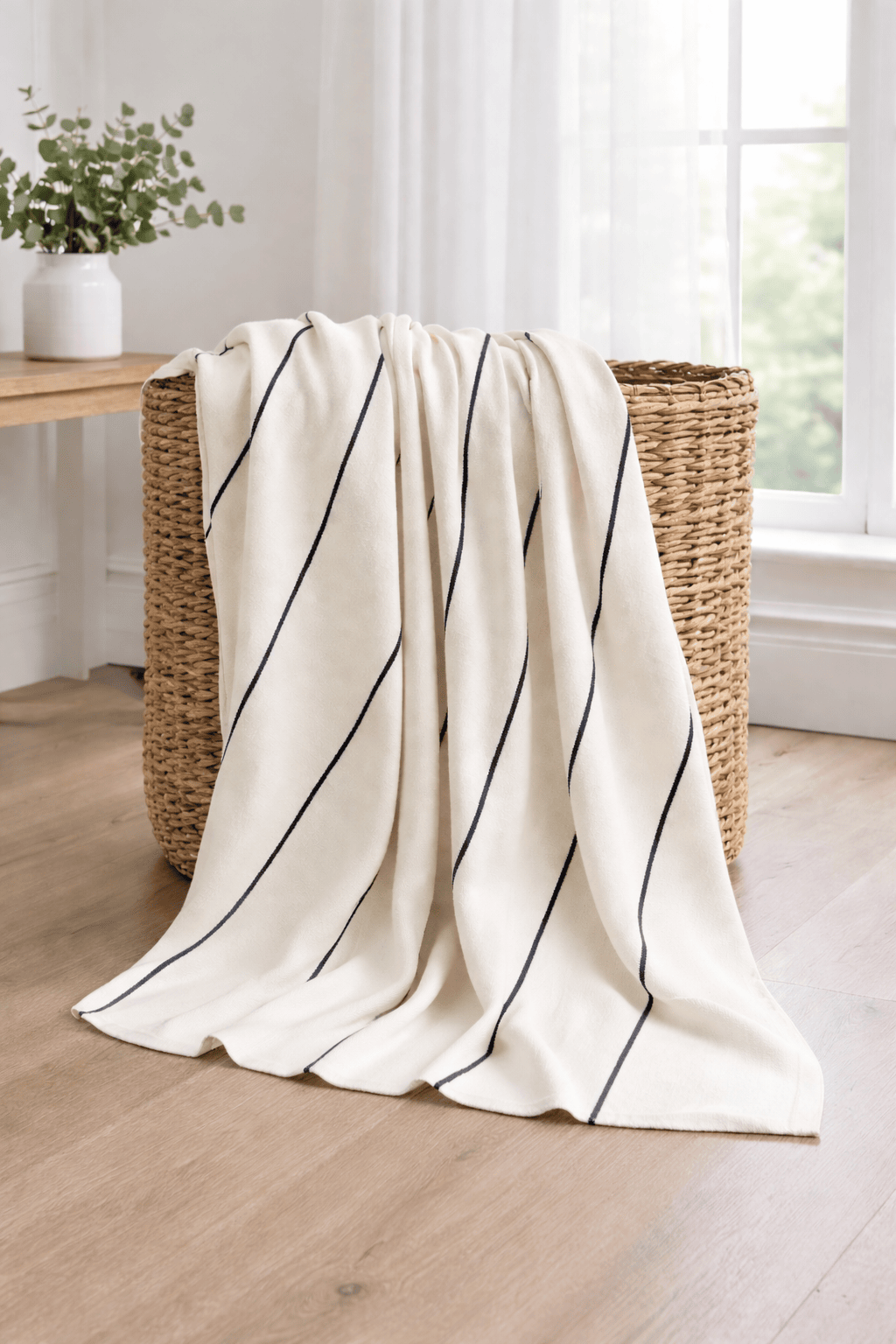 White cotton throw blanket with black stripes draped over a woven basket on a wooden floor.