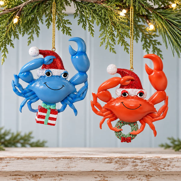 Decorative and whimsical red and blue crab with sparkly Santa hats carrying a wreath and present Christmas Ornaments hanging from a pine branch