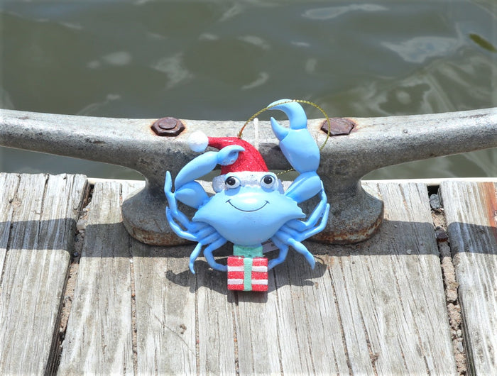 Blue crab nautical Christmas ornament wearing a red Santa hat and holding a gift box on a dock ready to get in the water